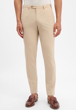 Beige Slim-Fit-Anzughose mit vorderem Knopf und Reißverschluss, getragen mit braunen Tassel-Loafers an einer stehenden Person.