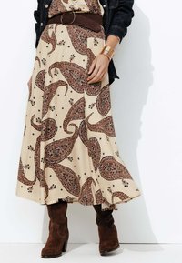 Robe maxi à motif paisley crème avec une ceinture et des accents marron. Le mannequin porte des bottines en daim marron. Une veste en denim est visible en arrière-plan.