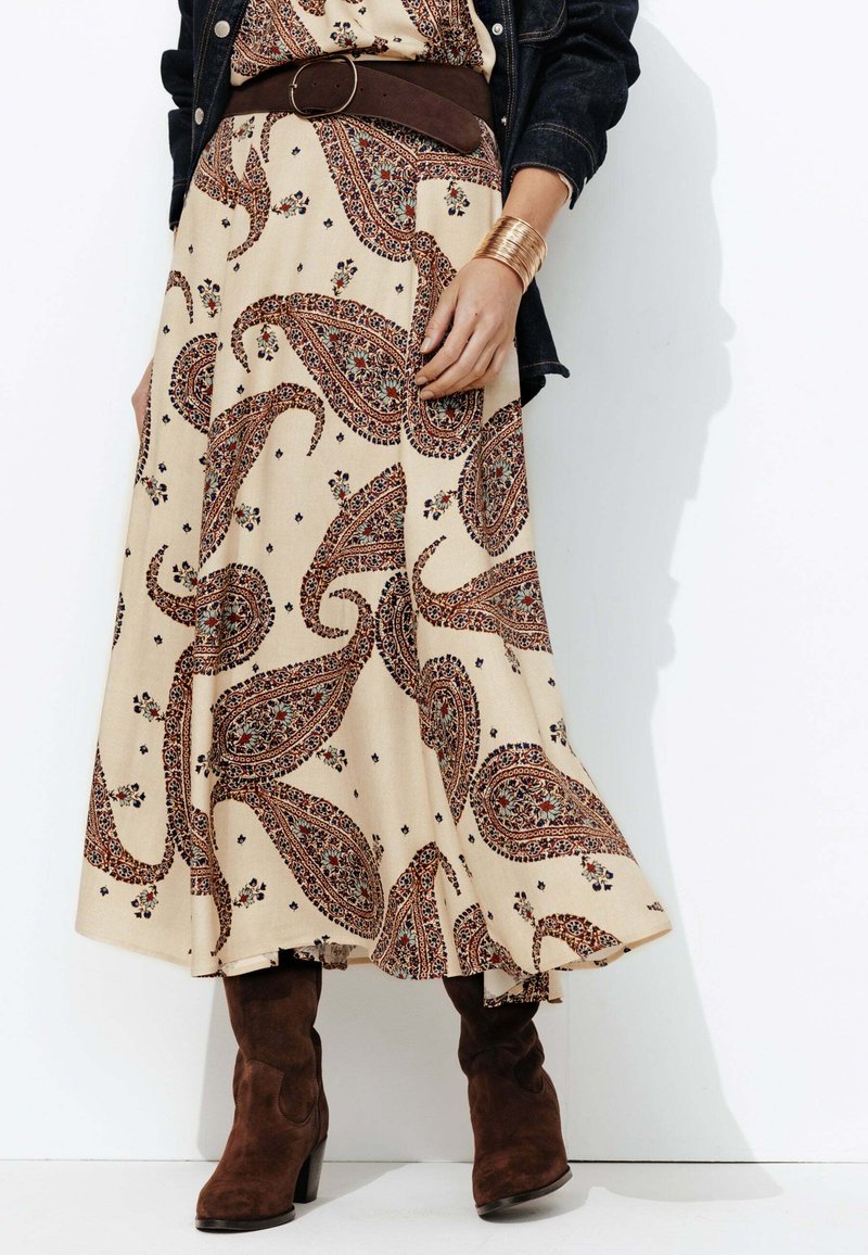Robe maxi à motif paisley crème avec une ceinture et des accents marron. Le mannequin porte des bottines en daim marron. Une veste en denim est visible en arrière-plan.