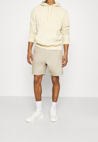 Man wearing light beige hoodie, matching beige shorts, white socks, and white sneakers standing against a plain white background.