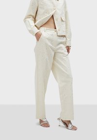 Crème-kleurig pak met een textuurafwerking, voorzien van metalen accenten. De jas heeft een cropped ontwerp en wordt gedragen met wijde broekspijpen en zilveren hakken.
