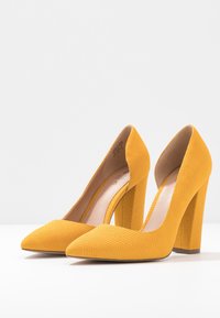 Chaussures à talons hauts jaunes avec un motif texturé ressemblant à celui des reptiles, des pointes effilées et des talons larges en bloc. Intérieur dans une couleur douce et claire.