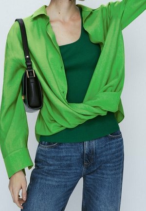 Person wearing a bright green button-up shirt over a dark green tank top, blue jeans, and carrying a small black shoulder bag.