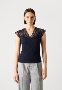 Haut sans manches en ribbed bleu marine avec un col en dentelle à volants et des manches en dentelle. Porté avec un pantalon plissé gris clair.