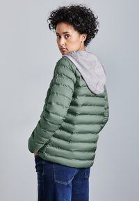 Grön puffjacka med en grå huva, med kviltad textur och ett logotypmärke på vänster arm. Buren över blå jeans.