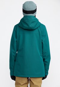 Veste en teck à capuche avec une texture lisse, dotée de poignets ajustables et de poches latérales. Portée sur un pantalon beige, accompagnée d'un bonnet en tricot.
