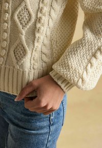 Krémový sveter s pleteným vzorom, s riasenými manžetami a voľným strihom, skombinovaný s modrými džínsami.
