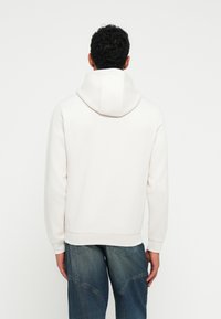 Sudadera blanca con capucha, de textura suave, corte relajado y puños acanalados, combinada con vaqueros azules con detalles de costura.