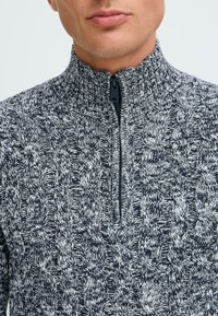 Pull en maille zippé en bleu marine et blanc, avec un motif en câble texturé et un col montant avec une fermeture éclair noire.