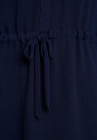 Robe en tissu bleu marine avec une taille à cordon. Texture douce et design fluide, avec un accent noué à la taille. Construction simple et élégante.