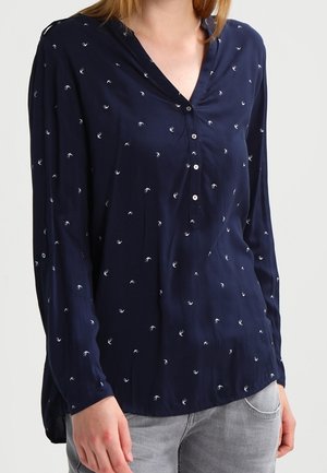 Femme portant un chemisier bleu marine avec de petits motifs d'oiseaux blancs, manches longues, et quatre boutons sur le devant, associé à un jean gris clair.