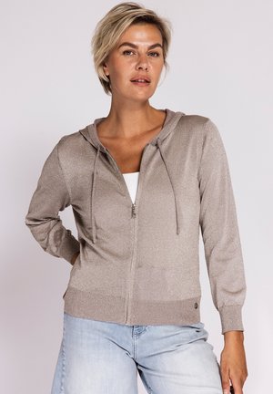 Frau mit kurzen blonden Haaren, die einen taupefarbenen Kapuzenpullover mit Reißverschluss über einem weißen Oberteil und hellblauen Jeans trägt, steht vor einem einfarbigen Hintergrund.
