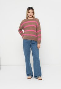 Brązowy sweter dzianinowy z różowym paskowanym wzorem, szerokim dekoltem, luźnym krojem, zestawiony z niebieskimi dzwonami i jasnymi butami.