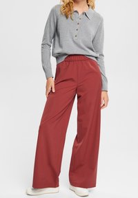 Top grigio a maniche lunghe con bottoni abbinato a pantaloni bordeaux a gamba larga, realizzati in un tessuto liscio, con vita elastica e sneakers bianche.