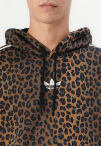 Primo piano di una persona che indossa un hoodie a stampa leopardata marrone con cordini neri e un logo Adidas bianco sul petto.