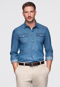Chemise en denim de couleur bleu clair, avec une patte de boutonnage sur le devant, deux poches poitrine et des manches retroussées, associée à un pantalon beige et une ceinture marron.
