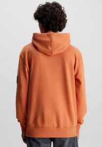 Orange huhoodtröja i mjukt tyg, med ribbade muddar och nederkant, samt en liten logotypmärke på ärmen, med en slät texturyta.