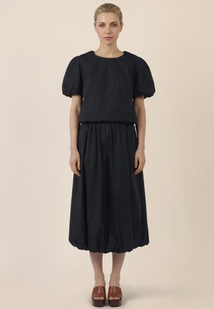 Woman with blonde hair in a black puff-sleeve top and matching midi skirt wearing brown platform sandals, standing against a beige background.