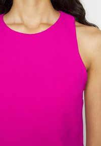 Vrouw die een mouwloos felroze topje met een ronde hals draagt, waarbij de nek, linkerschouder en een deel van het donkere haar zichtbaar zijn.