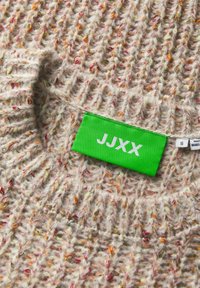 Stickad tröja i beige med flerfärgade fläckar, med en grön etikett med vit text "JJXX" och storlek "S" på halsen.