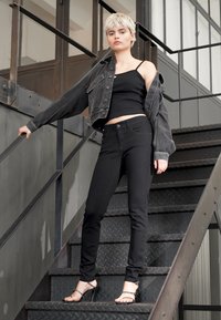 Fekete farmerdzseki fekete top felett és fekete, testhezálló farmernadrág. A modellen pántos fekete magassarkú cipő van. A háttér fém lépcsőket és ipari dizájnt mutat.