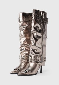 Cizme metalice bronz, până la genunchi, cu material texturat, vârfuri ascuțite, accente cu catarame și închidere cu fermoar. Prezintă curele cu ținte pentru detaliu.