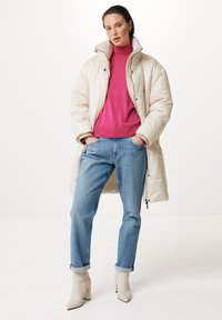 Abrigo acolchado beige con cuello alto, combinado con un suéter de cuello alto rosa brillante. Jeans de pierna recta en azul claro y botas de tobillo color crema completan el look.