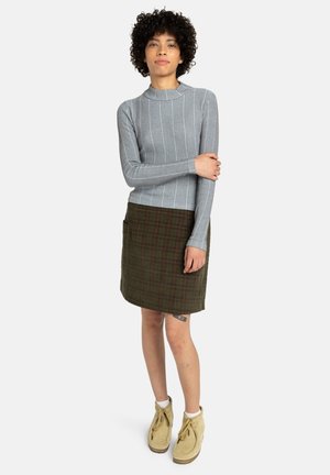 Grijze geribbelde lange mouwtop gecombineerd met een groen geruite rok. De outfit wordt aangevuld met beige enkellaarsjes met veters en een textiele textuur.