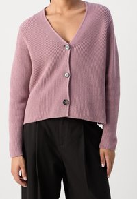 Różowy prążkowany kardigan z dekoltem w serek, trzema dużymi guzikami i krótkim krojem, w zestawieniu z czarnymi luźnymi spodniami.