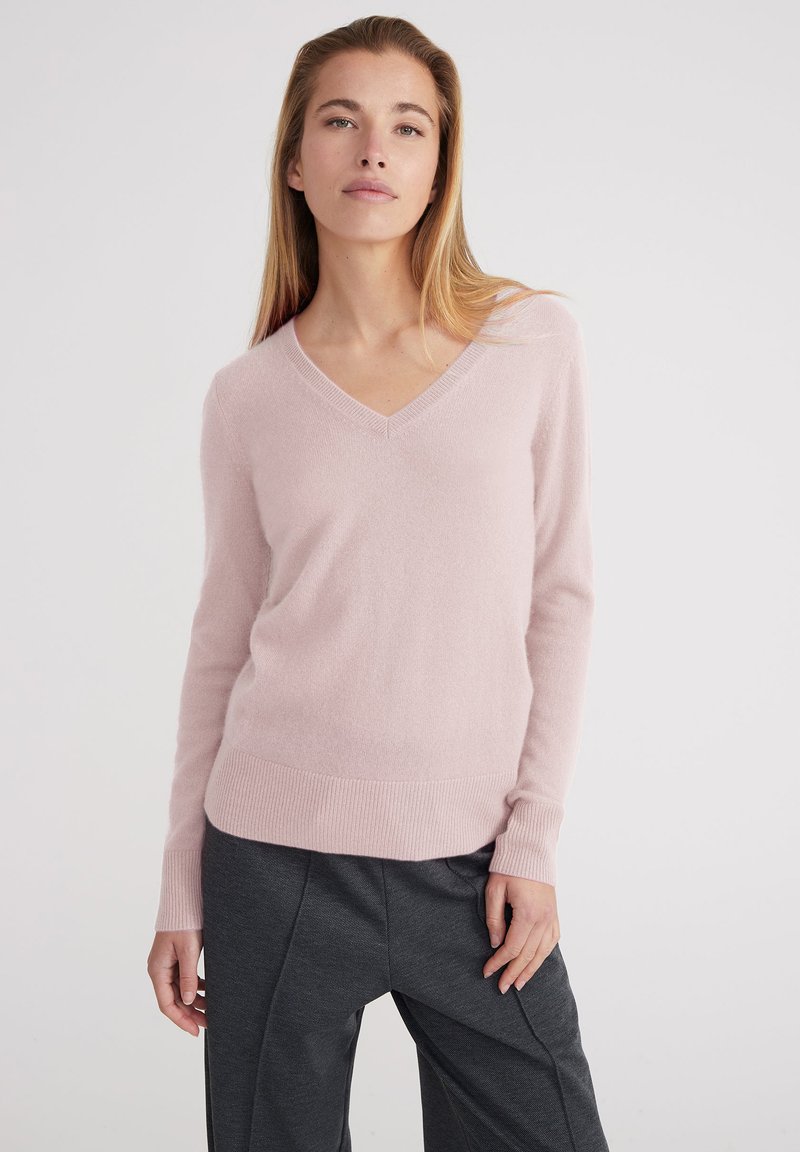 Donna con un maglione rosa chiaro a scollo a V e pantaloni grigio scuro, in piedi con un'espressione neutra contro uno sfondo chiaro e semplice.