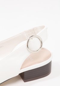 Sandale en cuir blanc avec un accent en anneau argenté rond et un petit talon en bois marron. Le design comprend un style à lanière pour un ajustement sécurisé.