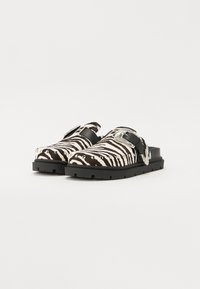 Zebra-print clogs with a black rubber sole, featuring a black strap and silver buckle accents. Textured upper with stud details.