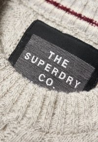 Szara etykieta z tkaniny jodełkowej na kremowym dzianym ubraniu, z białym wyszywanym napisem "THE SUPERDRY CO." oraz czerwonymi akcentującymi paskami powyżej.