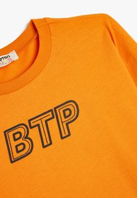 Oranssi puuvillapaita, jossa on musta, rohkea "BTP" -grafiikka edessä. Pyöreä kaula-aukko, sileä pinta ja lyhyet hihat.