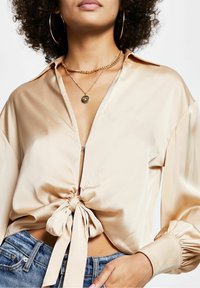 Kvinna i beige satinblus knuten fram med lång ärm, ihop med blå jeans och guldhalsband och hoop-örhängen.