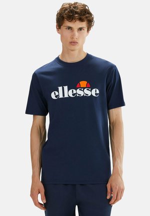 Young man with curly hair wearing a navy blue t-shirt with a white and orange ellesse logo, standing against a plain light background.