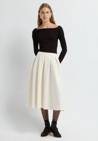 Femme portant un haut noir ajusté décolleté, une jupe midi plissée crème, des collants noirs transparents, des chaussettes sombres et des chaussures à lacets marron.