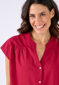 Blusa marrone con chiusura a bottoni, maniche corte e dettaglio arricciato intorno al collo, realizzata in un tessuto morbido.