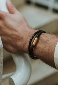 Musthave mustriviga nahast käevõru, millel on tsilindriline messingist lukk, kantud randmel heledal käsipuu taustal.