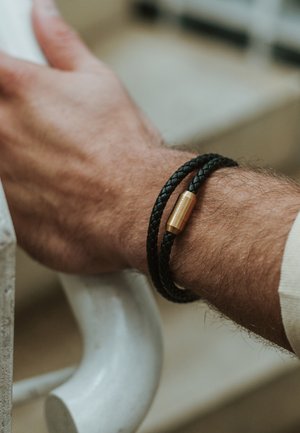 Black braided leather bracelet with a cylindrical brass clasp, worn on a wrist against a light-colored railing background.