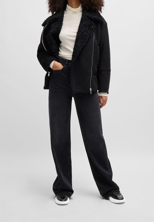 Personne portant une veste en peau lainée noire sur un col roulé blanc, un jean noir à jambes larges et des baskets noires et blanches, debout avec la main dans la poche.