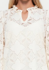 Vrouw in een witte kantblouse met bloemenpatronen en een geplooide kraag, die de bovenste torso en nek laat zien.