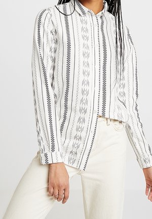 Femme portant une chemise blanche à manches longues avec des motifs verticaux noirs et gris, associée à un pantalon écru.