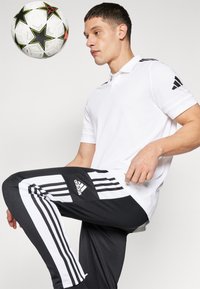 T-shirt polo blanc avec des accents noirs ; pantalon de sport noir avec des rayures blanches et le logo Adidas. Un ballon de soccer est en l'air au-dessus.