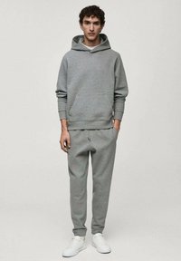 Felpa grigia con cappuccio e pantaloni della tuta coordinati, realizzati in tessuto morbido, dotati di tasca a marsupio, vita regolabile con cordino e polsini elasticizzati. Sneakers bianche.
