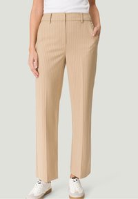 Pantalon beige à fines rayures avec une coupe droite, doté d'un devant plat, de poches latérales et d'une ceinture. Porté avec des baskets blanches.