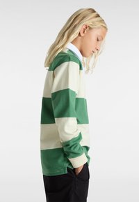 Een rugbyshirt met lange mouwen, voorzien van afwisselende groene en crème strepen, gemaakt van katoen, met een kraag en een comfortabele pasvorm, gedragen met zwarte broek.