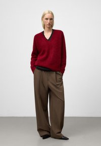 Czerwony sweter z wełny z głębokim dekoltem w kształcie V, w połączeniu z luźnymi brązowymi spodniami. Ubranie wyróżnia się teksturowanym wykończeniem oraz czarnymi akcentami.