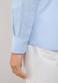 Camisa de manga larga azul claro con puños abotonados, que presenta una textura de tejido suave y dos botones en el borde de la manga.