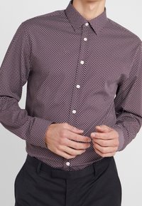 Camicia maniche lunghe di un profondo bordeaux con un motivo geometrico di diamanti bianchi, chiusura frontale con bottoni, colletto a punta e tessuto texturizzato.